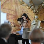 Eine Frau an der Geige und ein Mann am Keyboard spielen vor Publikum